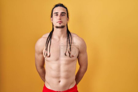 Téléchargez les photos : Hispanic man with long hair standing shirtless over yellow background relaxed with serious expression on face. simple and natural looking at the camera. - en image libre de droit