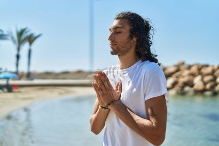Foto de Young hispanic man doing yoga exercise standing at beach - Imagen libre de derechos