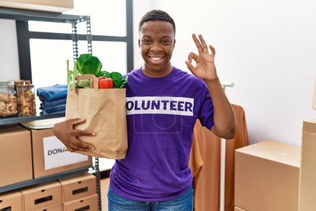 Téléchargez les photos : Young african volunteer man holding groceries doing ok sign with fingers, smiling friendly gesturing excellent symbol - en image libre de droit