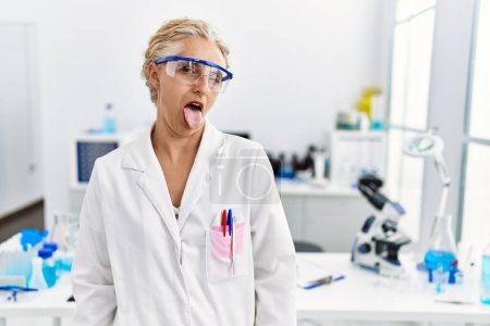 Téléchargez les photos : Femme blonde du moyen âge travaillant au laboratoire scientifique langue collante heureux avec une expression drôle. concept d'émotion. - en image libre de droit