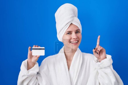 Photo for Blonde caucasian woman wearing bathrobe holding credit card surprised with an idea or question pointing finger with happy face, number one - Royalty Free Image