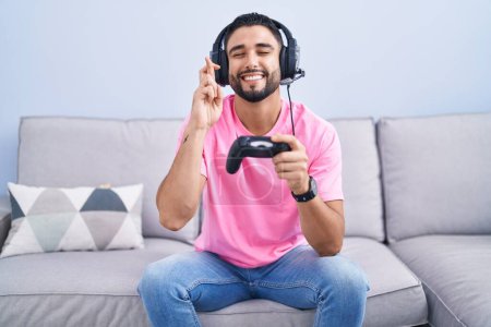 Photo for Hispanic young man playing video game holding controller sitting on the sofa gesturing finger crossed smiling with hope and eyes closed. luck and superstitious concept. - Royalty Free Image