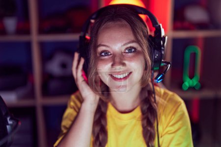Téléchargez les photos : Jeune femme blonde streamer souriant confiant assis sur la table à la salle de jeux - en image libre de droit