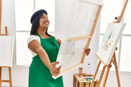 Foto de Mujer morena hispana sosteniendo lienzo orgulloso de pintar en estudio de arte - Imagen libre de derechos