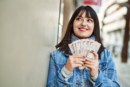 Photo for Young brunette woman holding arabia saudi riyal banknotes leaning on the wall - Royalty Free Image