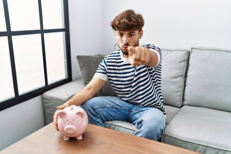 Téléchargez les photos : Young arab man holding piggy bank pointing with finger to the camera and to you, confident gesture looking serious - en image libre de droit