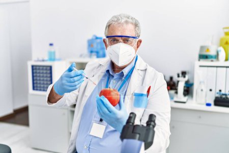 Foto de Hombre de pelo gris de mediana edad con uniforme científico con jeringa y manzana en el laboratorio - Imagen libre de derechos