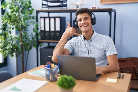 Téléchargez les photos : Jeune homme hispanique travaillant au bureau portant des écouteurs souriant faire geste de téléphone avec la main et les doigts comme parler au téléphone. communiquer les concepts. - en image libre de droit