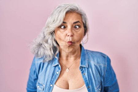 Téléchargez les photos : Femme d'âge moyen avec les cheveux gris debout sur fond rose faisant visage de poisson avec des lèvres, geste fou et comique. expression drôle. - en image libre de droit