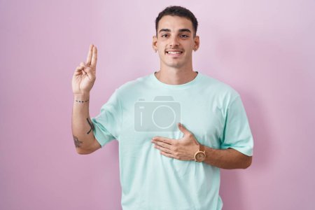 Téléchargez les photos : Bel homme hispanique debout sur fond rose souriant jurant avec la main sur la poitrine et les doigts vers le haut, faisant un serment de fidélité promesse - en image libre de droit