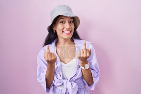 Foto de Young hispanic woman standing over pink background wearing hat showing middle finger doing fuck you bad expression, provocation and rude attitude. screaming excited - Imagen libre de derechos