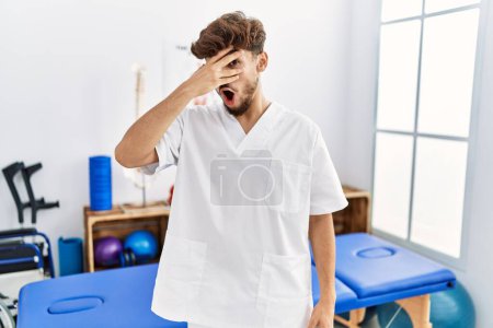 Photo for Young arab man working at pain recovery clinic peeking in shock covering face and eyes with hand, looking through fingers with embarrassed expression. - Royalty Free Image