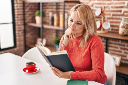 Téléchargez les photos : Jeune femme blonde lecture livre et boire du café à la maison - en image libre de droit