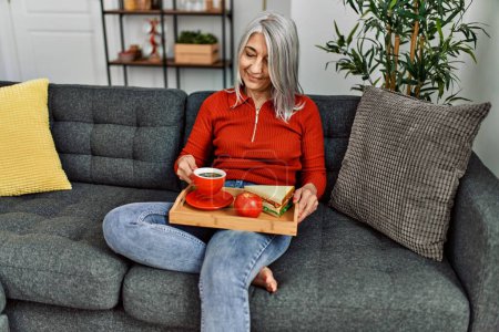 Téléchargez les photos : Femme aux cheveux gris d'âge moyen prenant le petit déjeuner assis sur le canapé à la maison - en image libre de droit