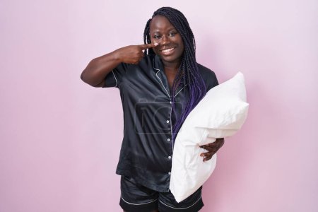 Téléchargez les photos : Jeune femme africaine portant un pyjama étreignant l'oreiller pointant du doigt vers le visage et le nez, souriante et joyeuse. concept de beauté - en image libre de droit