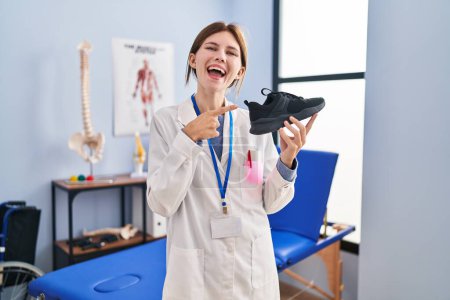 Photo for Young physiotherapist woman holding sneakers smiling happy pointing with hand and finger - Royalty Free Image