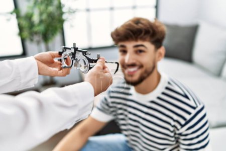 Téléchargez les photos : Jeune homme arabe souriant confiant recevant des lunettes optométriste à la maison - en image libre de droit