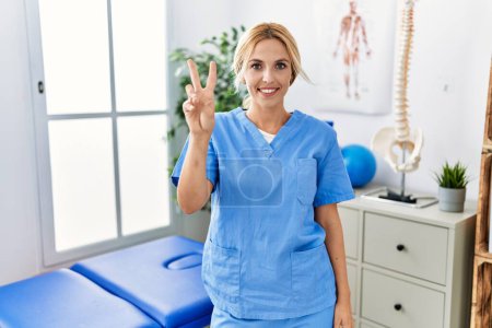 Photo for Beautiful blonde physiotherapist woman working at pain recovery clinic showing and pointing up with fingers number two while smiling confident and happy. - Royalty Free Image