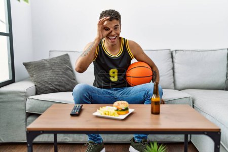 Photo for Young handsome hispanic man holding basketball ball cheering tv game smiling happy doing ok sign with hand on eye looking through fingers - Royalty Free Image