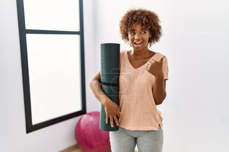 Téléchargez les photos : Young african american woman holding yoga mat at pilates room pointing thumb up to the side smiling happy with open mouth - en image libre de droit