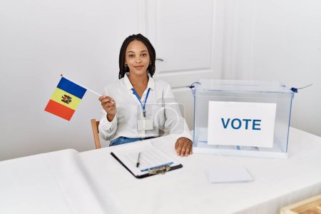Téléchargez les photos : Jeune femme afro-américaine souriante confiante tenant le drapeau moldova travaillant au collège électoral - en image libre de droit