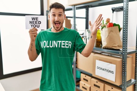 Téléchargez les photos : Jeune homme hispanique portant un t-shirt volontaire tenant nous avons besoin de vous bannière célébrant la victoire avec sourire heureux et expression gagnante avec les mains levées - en image libre de droit