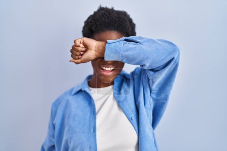 Foto de Mujer afroamericana de pie sobre fondo azul cubriendo los ojos con el brazo sonriendo alegre y divertido. concepto ciego. - Imagen libre de derechos