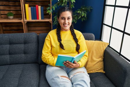 Téléchargez les photos : Jeune femme hispanique lecture livre assis sur le canapé à la maison - en image libre de droit