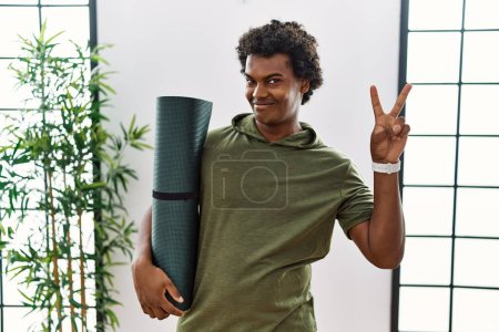 Téléchargez les photos : Homme africain aux cheveux bouclés tenant tapis de yoga en studio souriant avec un sourire heureux clin d'oeil à la caméra faisant signe de victoire avec les doigts. numéro deux. - en image libre de droit