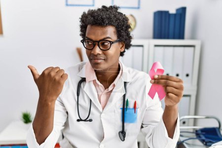 Photo for African doctor man holding pink cancer ribbon pointing thumb up to the side smiling happy with open mouth - Royalty Free Image