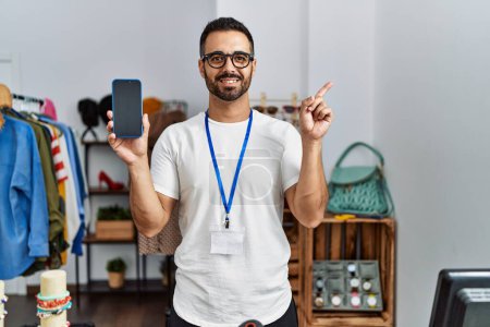 Foto de Joven hombre hispano con barba trabajando como gerente en la boutique minorista sosteniendo el teléfono inteligente sonriendo feliz señalando con la mano y el dedo hacia un lado - Imagen libre de derechos