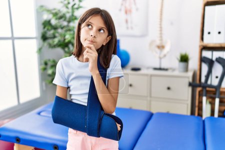 Photo for Young hispanic girl wearing arm on sling at rehabilitation clinic serious face thinking about question with hand on chin, thoughtful about confusing idea - Royalty Free Image