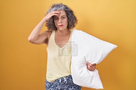 Foto de Mujer de mediana edad con el pelo gris usando pijama abrazando almohada preocupada y estresada por un problema con la mano en la frente, nerviosa y ansiosa por la crisis - Imagen libre de derechos