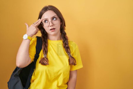 Foto de Joven mujer caucásica usando mochila de estudiante sobre fondo amarillo disparando y matándose señalando la mano y los dedos a la cabeza como arma, gesto suicida. - Imagen libre de derechos