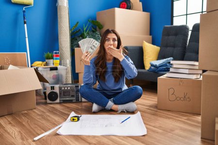 Photo for Young brunette woman sitting on the floor at new home holding money covering mouth with hand, shocked and afraid for mistake. surprised expression - Royalty Free Image