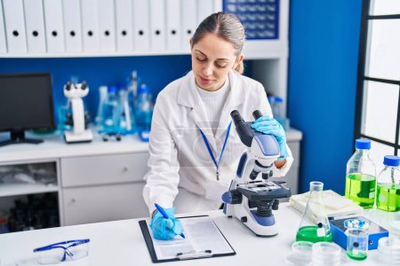 Téléchargez les photos : Jeune femme scientifique utilisant le microscope écrit sur le rapport au laboratoire - en image libre de droit