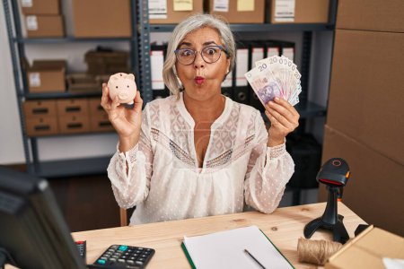 Photo for Middle age woman with grey hair working at small business ecommerce holding piggy bank and zloty making fish face with mouth and squinting eyes, crazy and comical. - Royalty Free Image