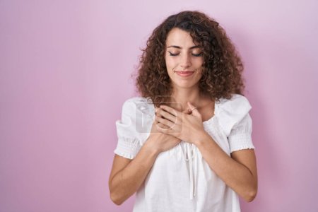 Téléchargez les photos : Hispanic woman with curly hair standing over pink background smiling with hands on chest with closed eyes and grateful gesture on face. health concept. - en image libre de droit