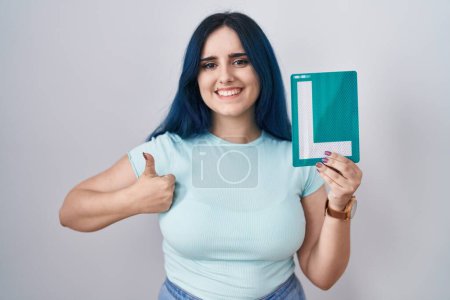 Foto de Young modern girl with blue hair holding l sign for new driver smiling happy and positive, thumb up doing excellent and approval sign - Imagen libre de derechos