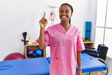 Téléchargez les photos : Jeune femme afro-américaine travaillant à la clinique de récupération de la douleur montrant et pointant du doigt le numéro un tout en souriant confiant et heureux. - en image libre de droit