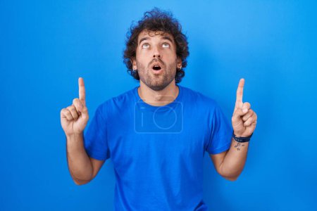 Photo for Hispanic young man standing over blue background amazed and surprised looking up and pointing with fingers and raised arms. - Royalty Free Image