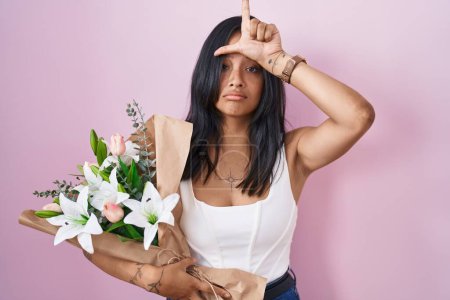 Photo for Brunette woman holding bouquet of white flowers making fun of people with fingers on forehead doing loser gesture mocking and insulting. - Royalty Free Image