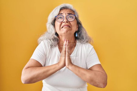 Photo for Middle age woman with grey hair standing over yellow background begging and praying with hands together with hope expression on face very emotional and worried. begging. - Royalty Free Image