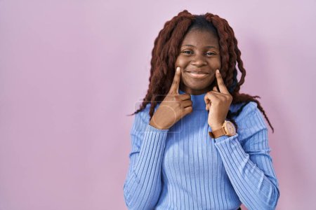 Foto de Mujer africana de pie sobre fondo rosa sonriendo con la boca abierta, los dedos señalando y forzando sonrisa alegre - Imagen libre de derechos