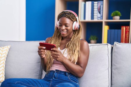 Téléchargez les photos : Femme afro-américaine regarder la vidéo sur smartphone assis sur le canapé à la maison - en image libre de droit