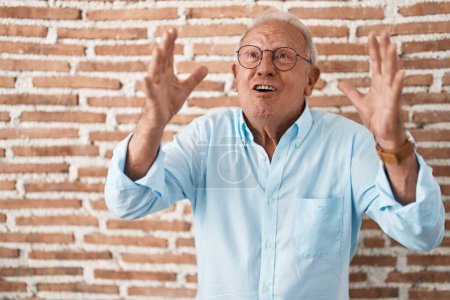 Foto de Hombre mayor con el pelo gris de pie sobre ladrillos pared loco y loco gritando y gritando con expresión agresiva y los brazos levantados. concepto de frustración. - Imagen libre de derechos
