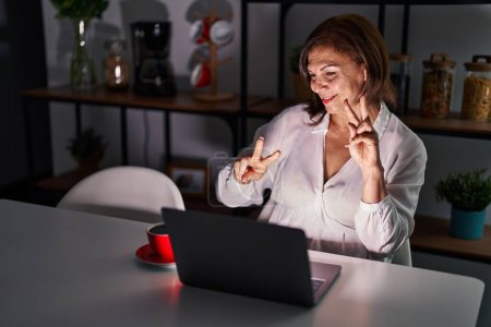 Téléchargez les photos : Femme hispanique du Moyen Age utilisant un ordinateur portable à la maison la nuit souriant en regardant la caméra montrant les doigts faisant signe de victoire. numéro deux. - en image libre de droit