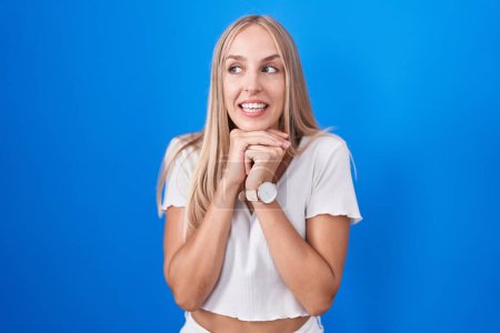 Foto de Young caucasian woman standing over blue background laughing nervous and excited with hands on chin looking to the side - Imagen libre de derechos
