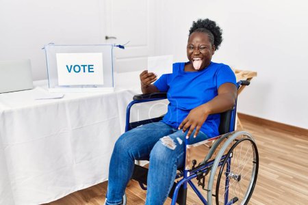 Foto de Mujer africana joven sentada en silla de ruedas votando poniendo sobre en las urnas la lengua que sobresale feliz con expresión divertida. concepto de emoción. - Imagen libre de derechos