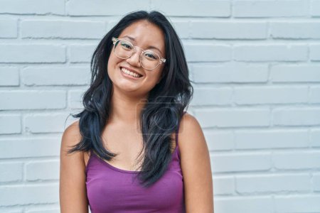 Foto de Asiática joven mujer de pie sobre ladrillos fondo buscando positivo y feliz de pie y sonriendo con una sonrisa confiada mostrando los dientes - Imagen libre de derechos